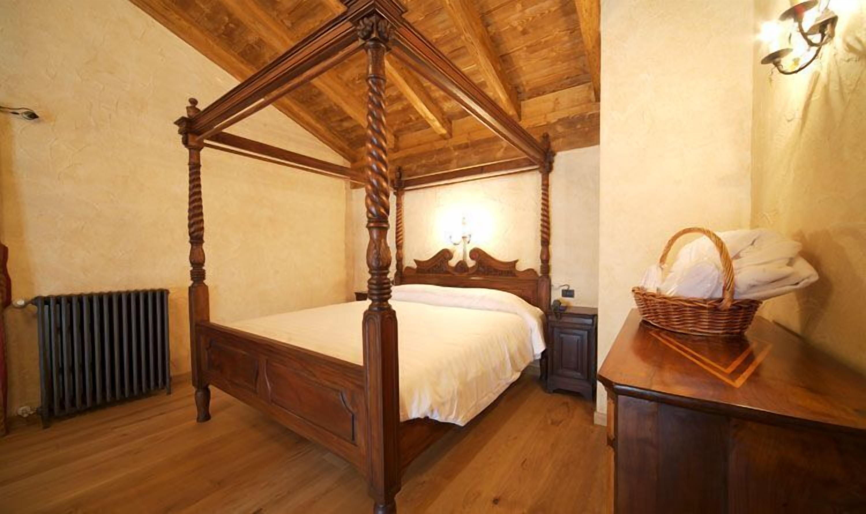 A four-poster bed with a wooden headboard, a nightstand, a wall-mounted lamp, and a wicker basket with towels.