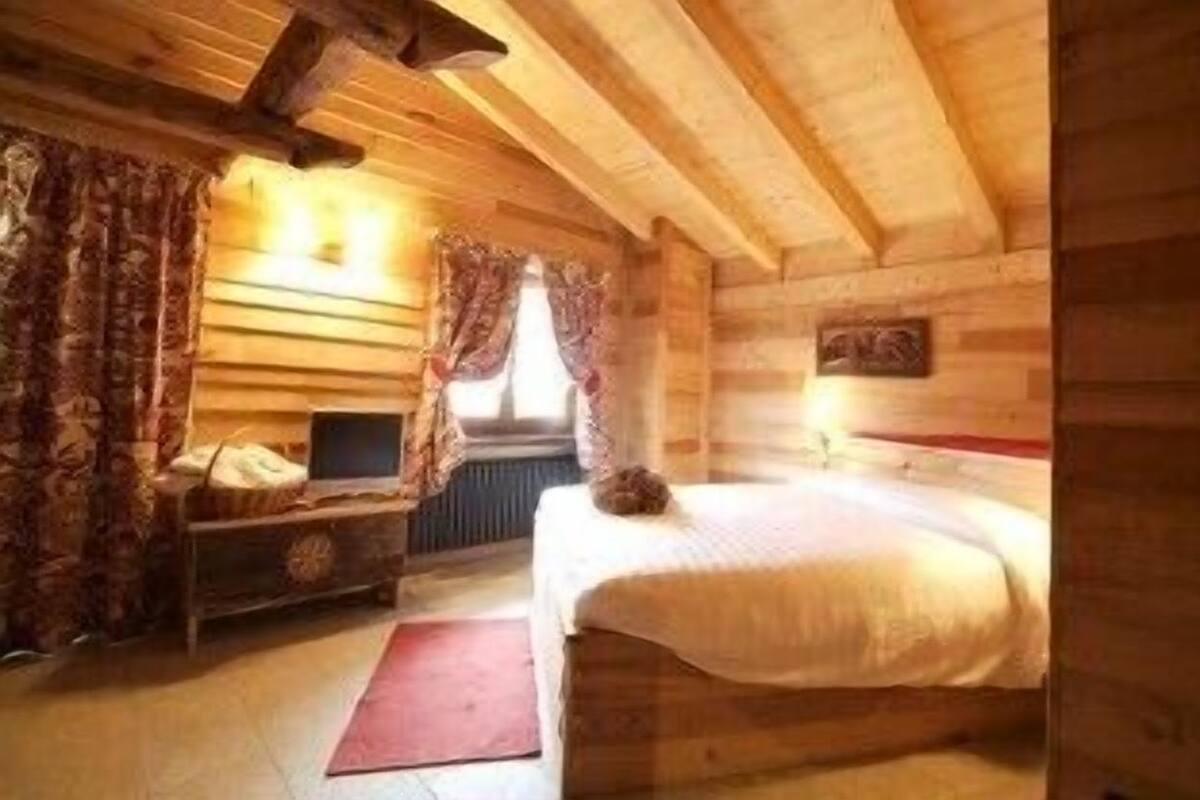 A wooden cabin bedroom with a bed, a dresser, a window with curtains, and a wooden ceiling.