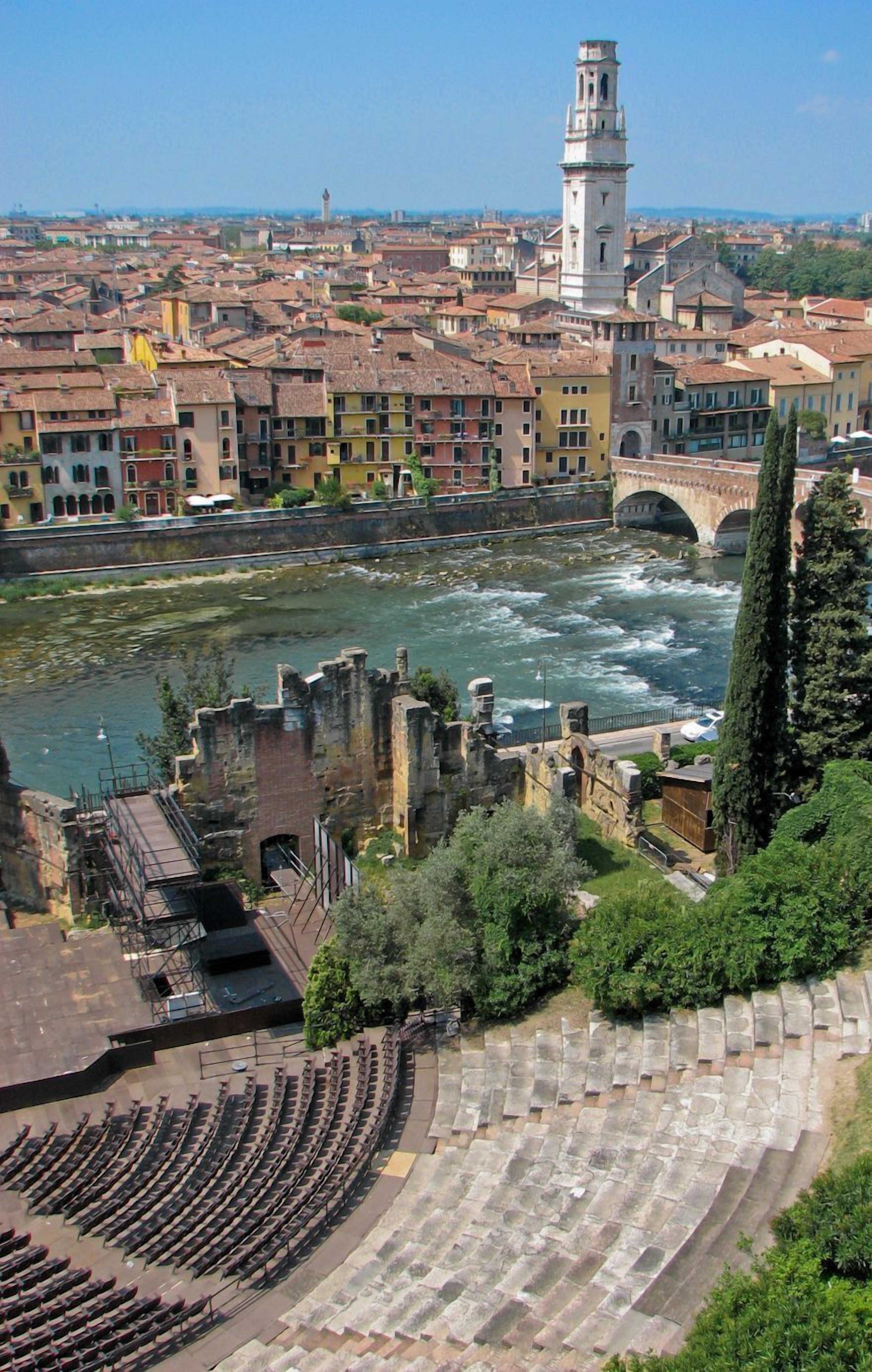 A historic theater with stone steps and rows of seating, overlooking a river and a cityscape with a prominent tower.