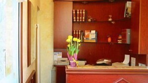 Un mostrador de recepción con detalles de madera, una placa identificativa, un jarrón con flores amarillas y una estantería con diversos objetos.