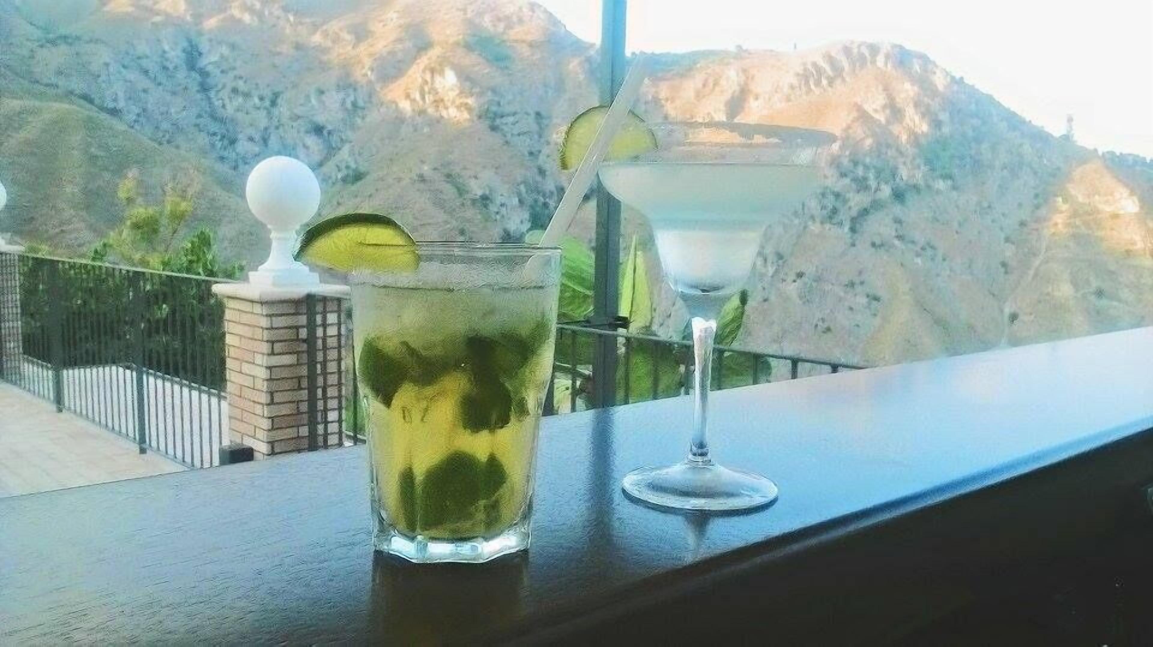 Two glasses of a green beverage with a lime slice on a balcony overlooking a mountainous landscape.