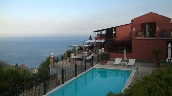 A poolside area with a view of the sea, a red building, and outdoor seating. - Il Casale di Caterina (Castelmola)