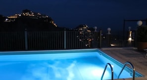 Une piscine avec un escalier en métal, entourée d’une clôture, et d’où l’on peut admirer une colline illuminée.