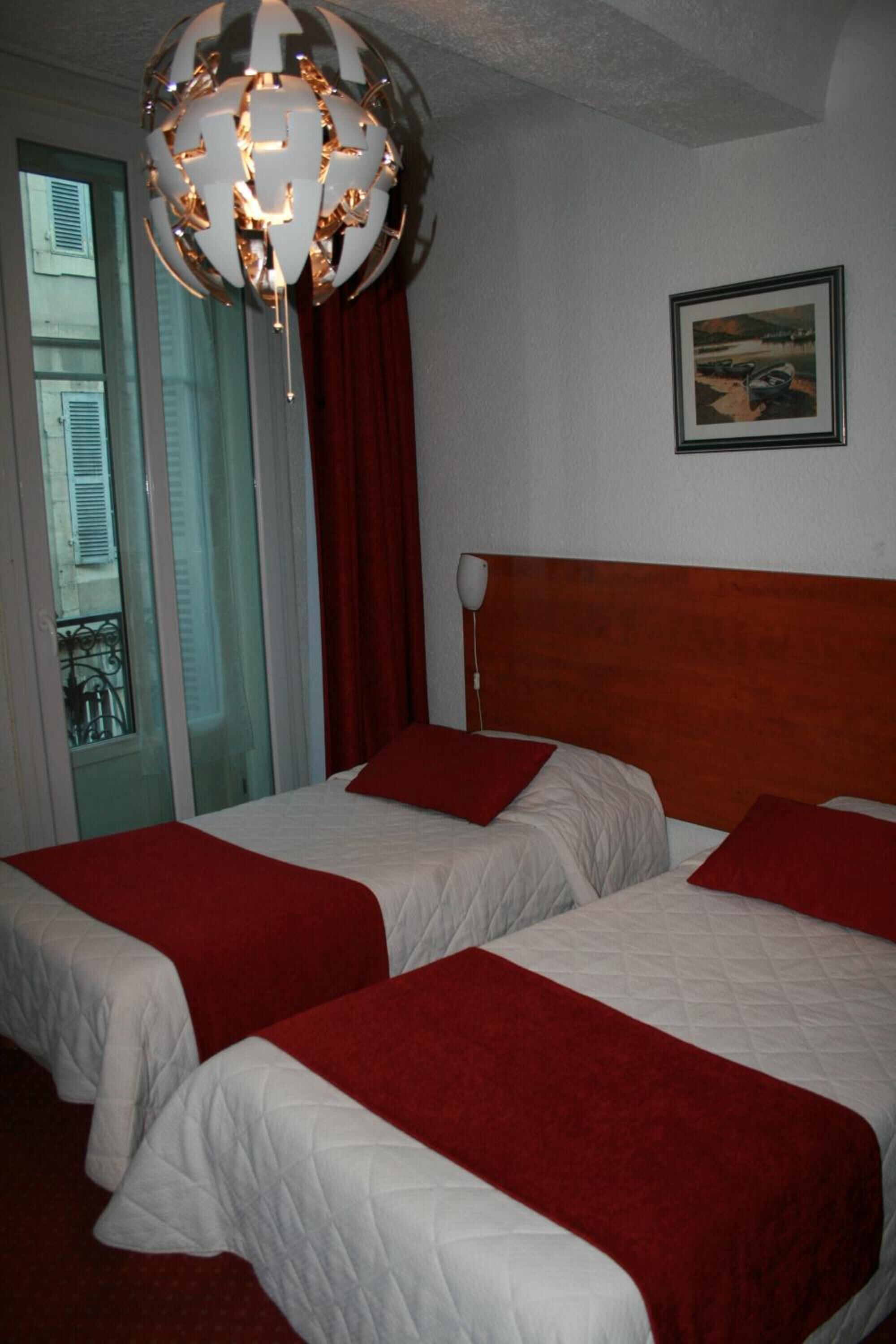 Una habitación de hotel con dos camas, un cabecero de madera, una cortina roja, una lámpara de araña con forma de globo y un cuadro en la pared.