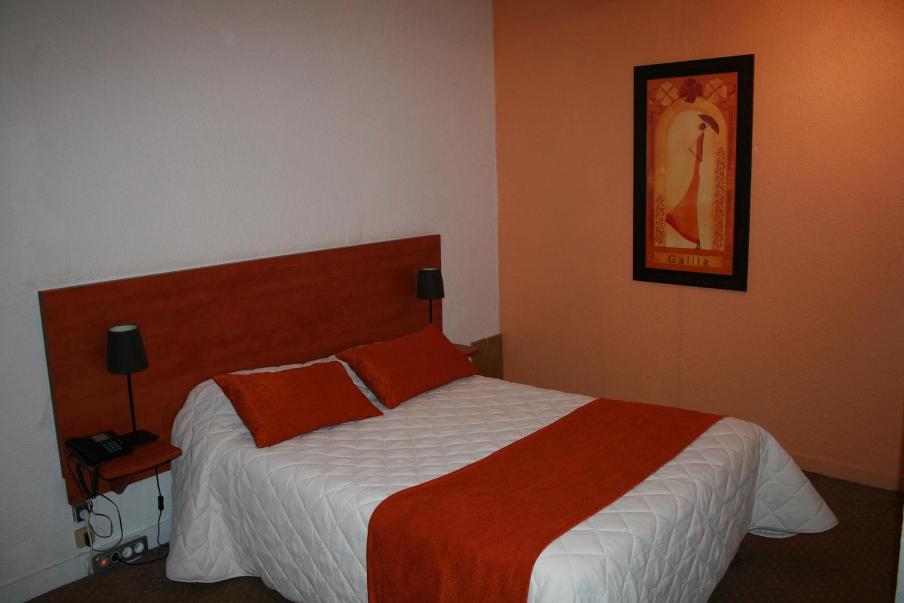 Una cama bien tendida con colcha blanca y almohadas rojas, un cabecero de madera, una mesita de noche con lámpara y un cuadro en la pared.