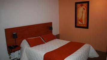 Una cama bien tendida con colcha blanca y almohadas rojas, un cabecero de madera, una mesita de noche con lámpara y un cuadro en la pared.