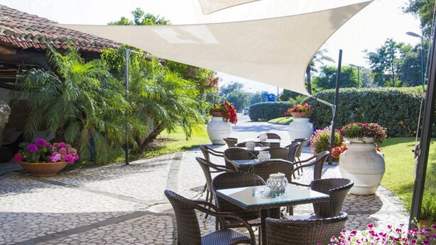 Un patio al aire libre con mobiliario de mimbre, plantas en macetas y un amplio paraguas que ofrece sombra.