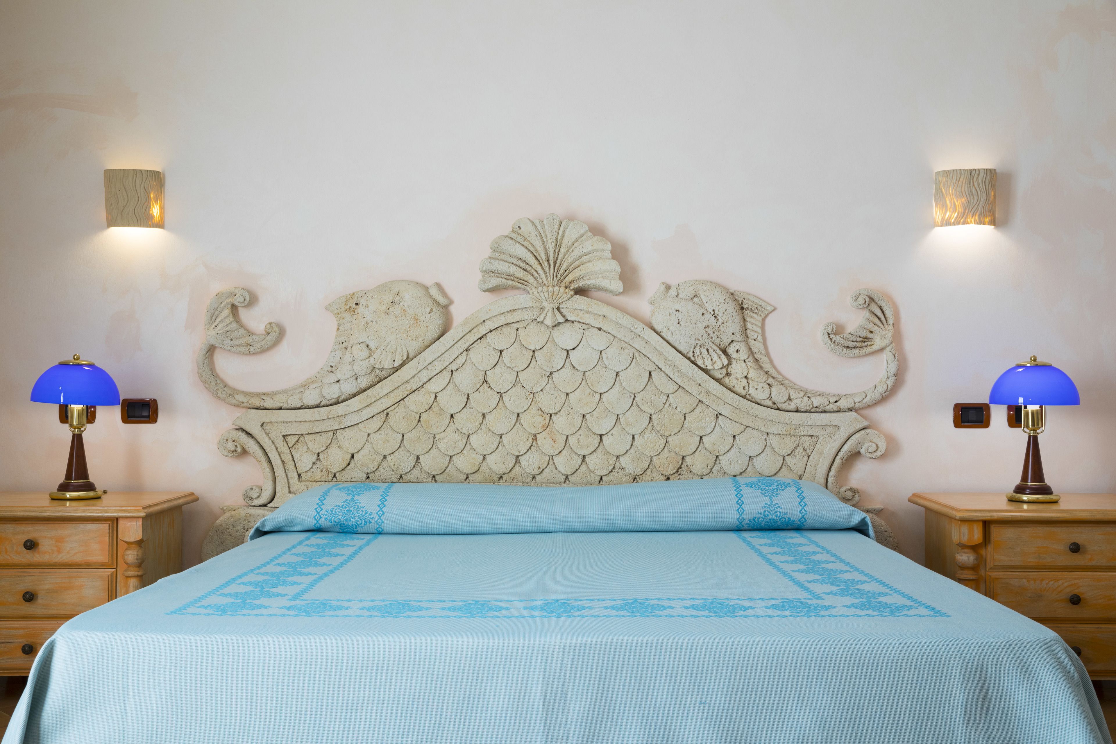 A bedroom with a decorative headboard, two bedside lamps, a bed with a blue bedspread, and a wooden nightstand.