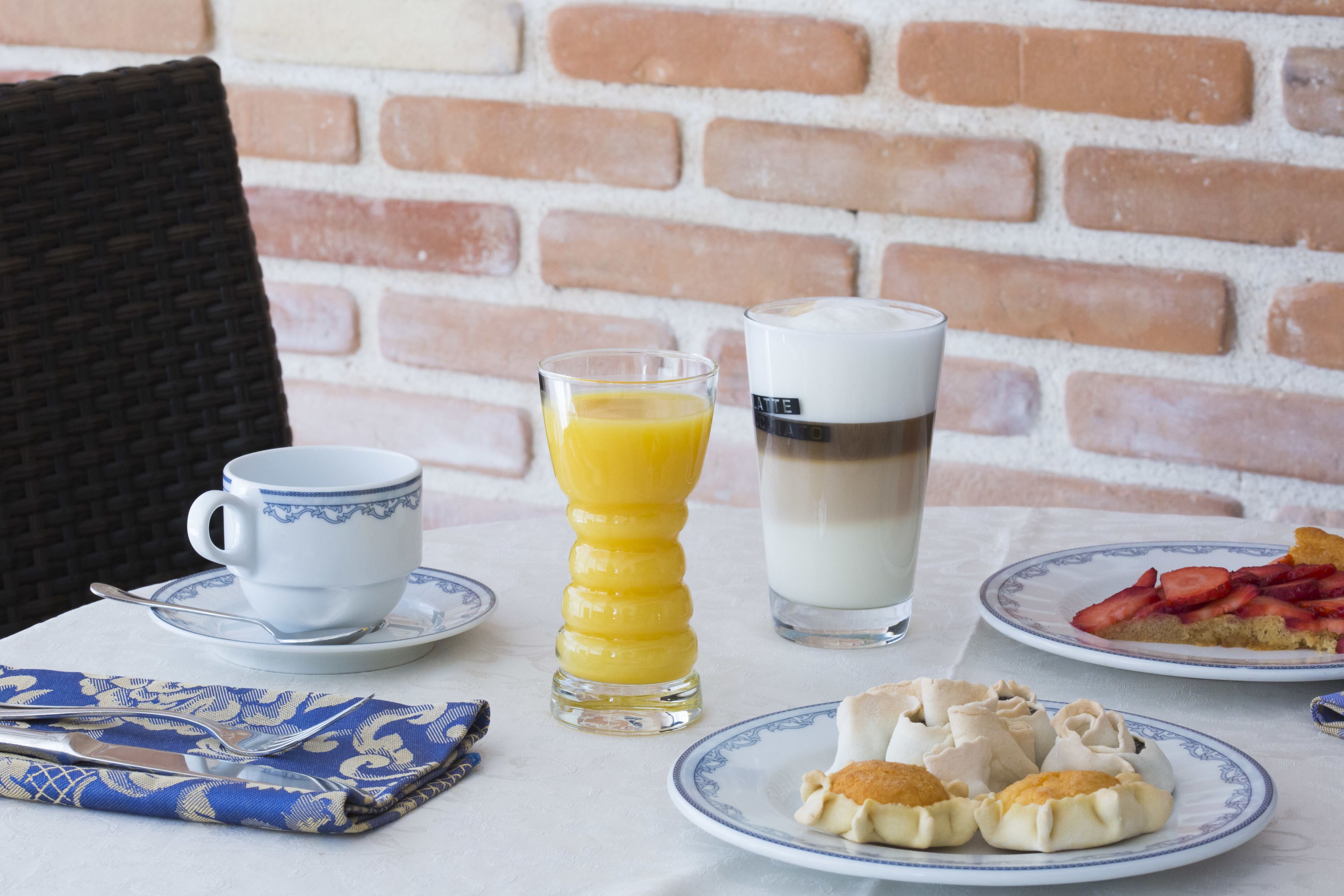Un desayuno con una taza, un vaso de zumo de naranja, un vaso de leche y platos con bollería y una porción de tarta.