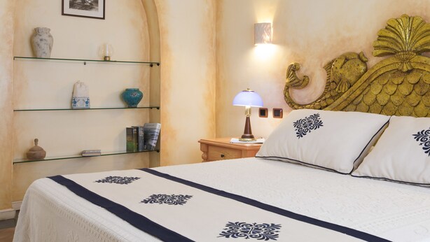 A neatly arranged hotel room with a bed, a nightstand, a lamp, and shelves with decorative items. - Hotel Poseidonia (Tortoli)