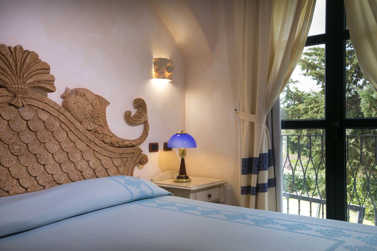 Un dormitorio con un cabecero decorativo, una mesita de noche con lámpara y vistas a árboles por la ventana.