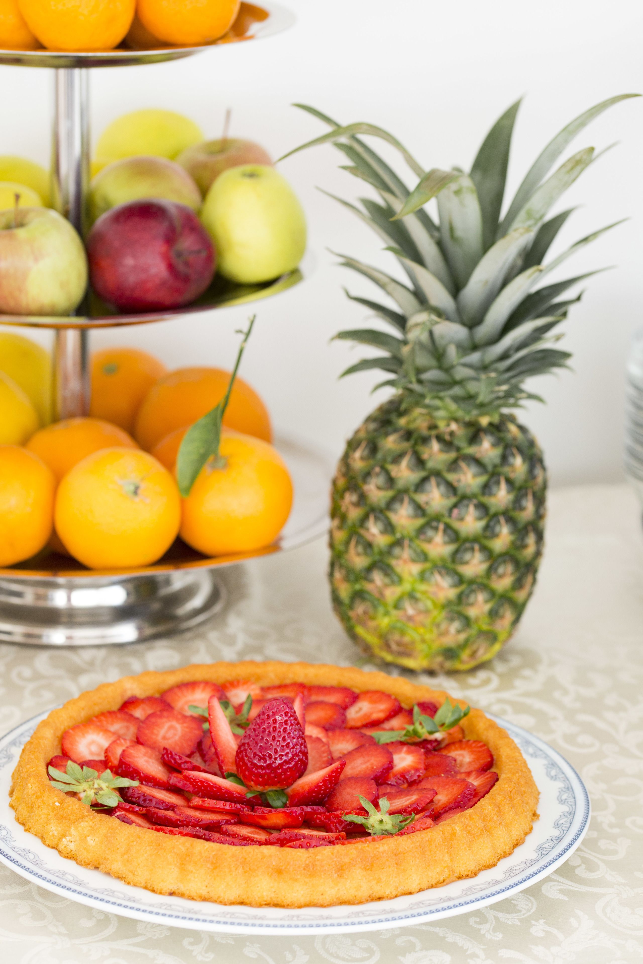 Una bandeja de frutas con naranjas, manzanas y una piña, junto con una tarta de fresas en un plato.
