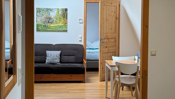 Una habitación con piso de madera, un sofá negro, una mesa de comedor con sillas, una puerta de madera y un cuadro en la pared.