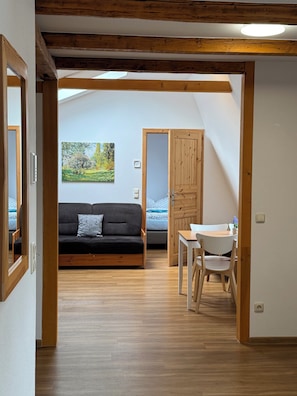 A room with a wooden floor, a black sofa, a dining table with chairs, a wooden door, and a painting on the wall.