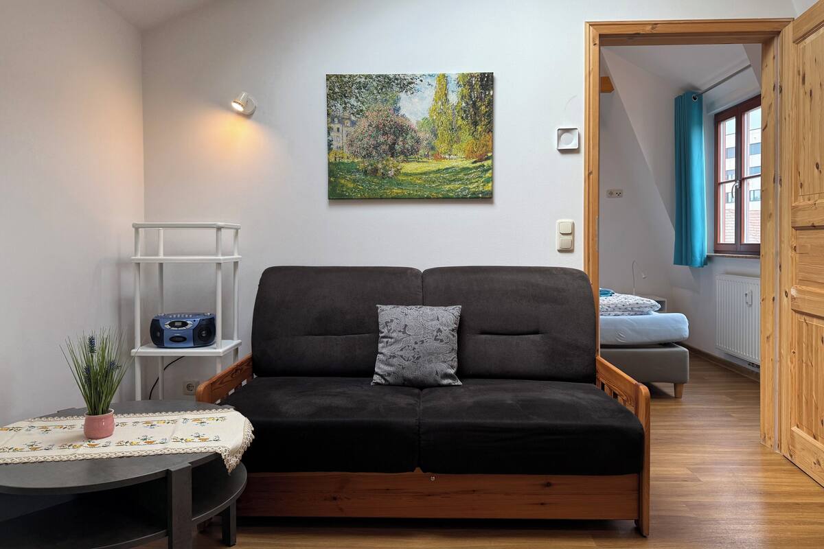 A modern living room with a black sofa, a small table with a lace tablecloth, a potted plant, a wall-mounted picture, and a wooden door leading to another room.