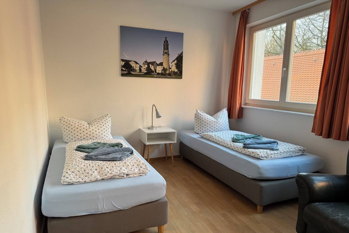 A room with two beds, a small table, a chair, and a window with curtains.