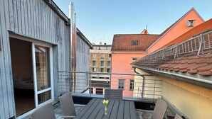 Terraza en la azotea con una mesa de madera y sillas de mimbre, una puerta de cristal que da a un espacio interior, y edificios al fondo.