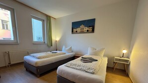 Una habitación con dos camas, un radiador, una ventana con cortinas, un cuadro en la pared y una mesita de noche con lámpara.