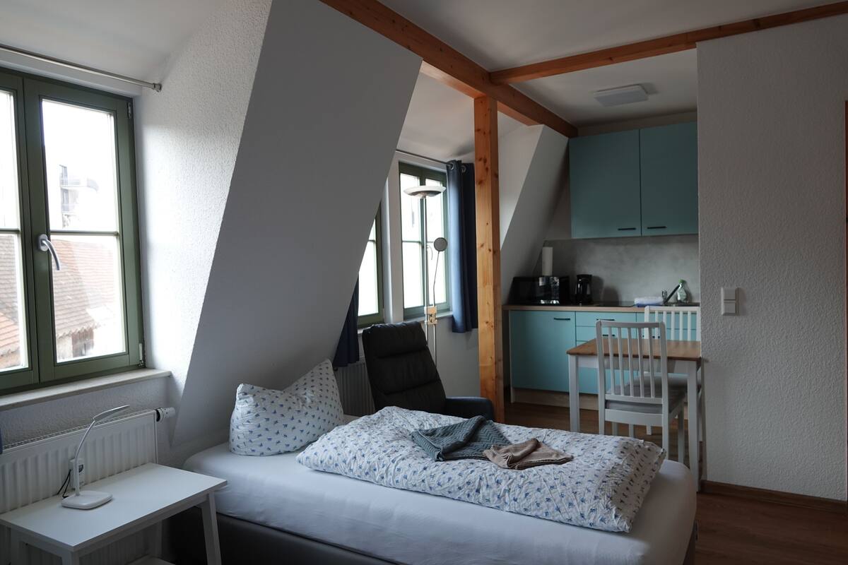 A bedroom with a bed, a desk, a chair, a window, and a kitchen area.