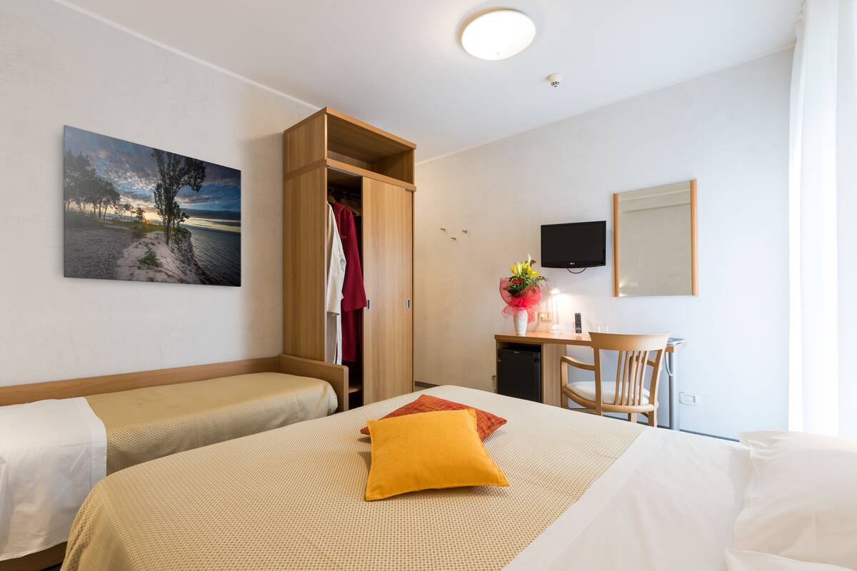 Une chambre d’hôtel avec deux lits, une armoire en bois, un bureau avec une chaise, une télévision et un tableau représentant un coucher de soleil.