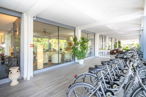 A modern hotel lobby with a row of bicycles, a large potted plant, and a glass wall offering a view of the outdoor seating area. - Hotel al Cigno (Lignano Sabbiadoro)