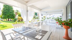 Un patio al aire libre con mobiliario de mimbre blanco, mesas de comedor ordenadas y una maceta con flores rojas.