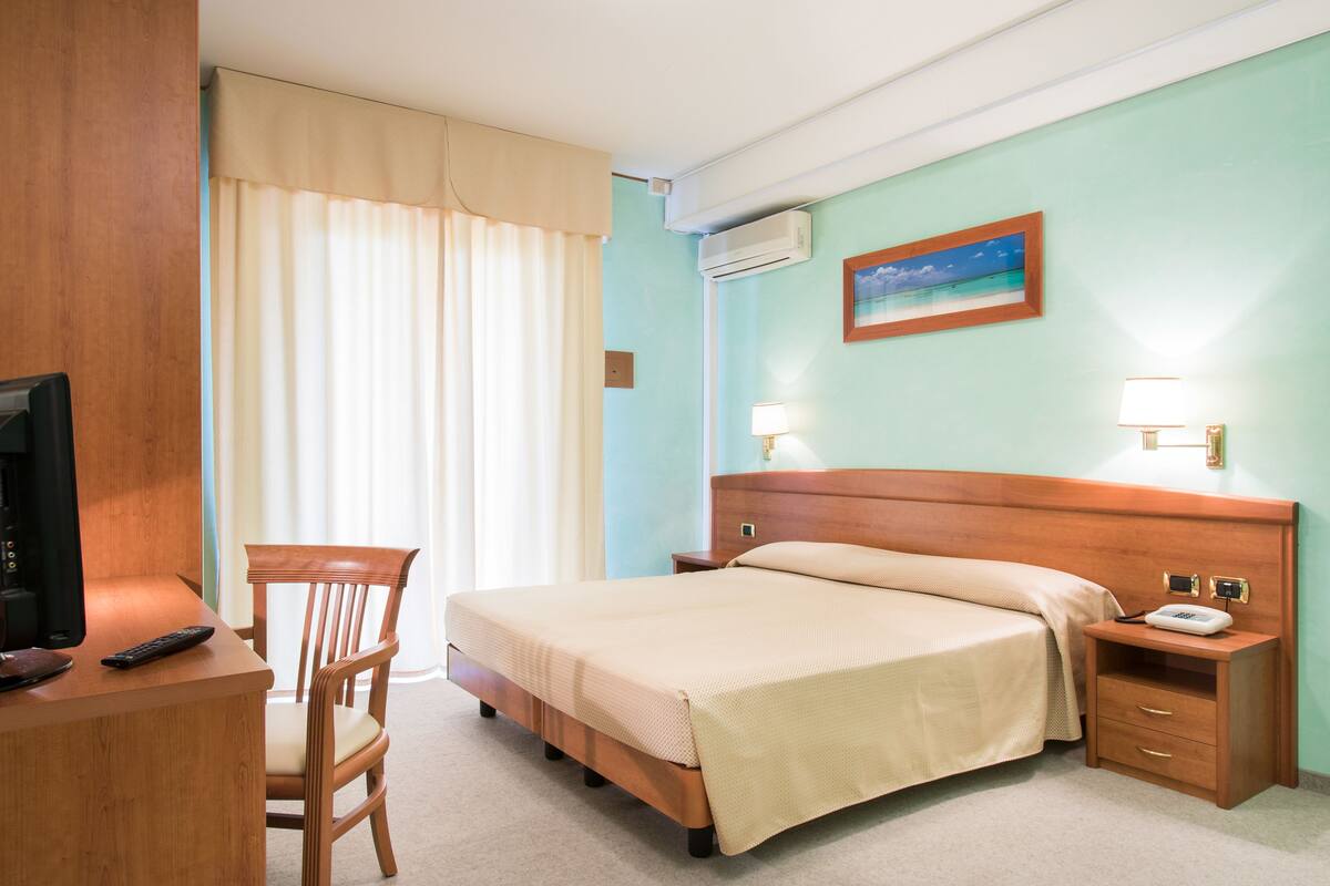 Une chambre d’hôtel comprenant un lit, un bureau avec une chaise, une télévision, une table de chevet avec une lampe et un appareil de climatisation.