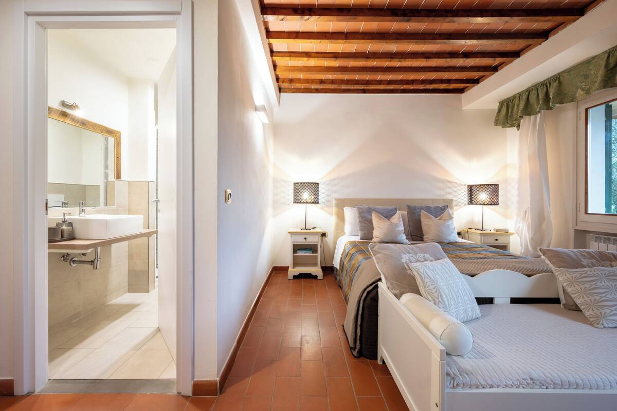 Een slaapkamer met een houten plafond, een bed met veel kussens, een nachtkastje en een raam met gordijnen.