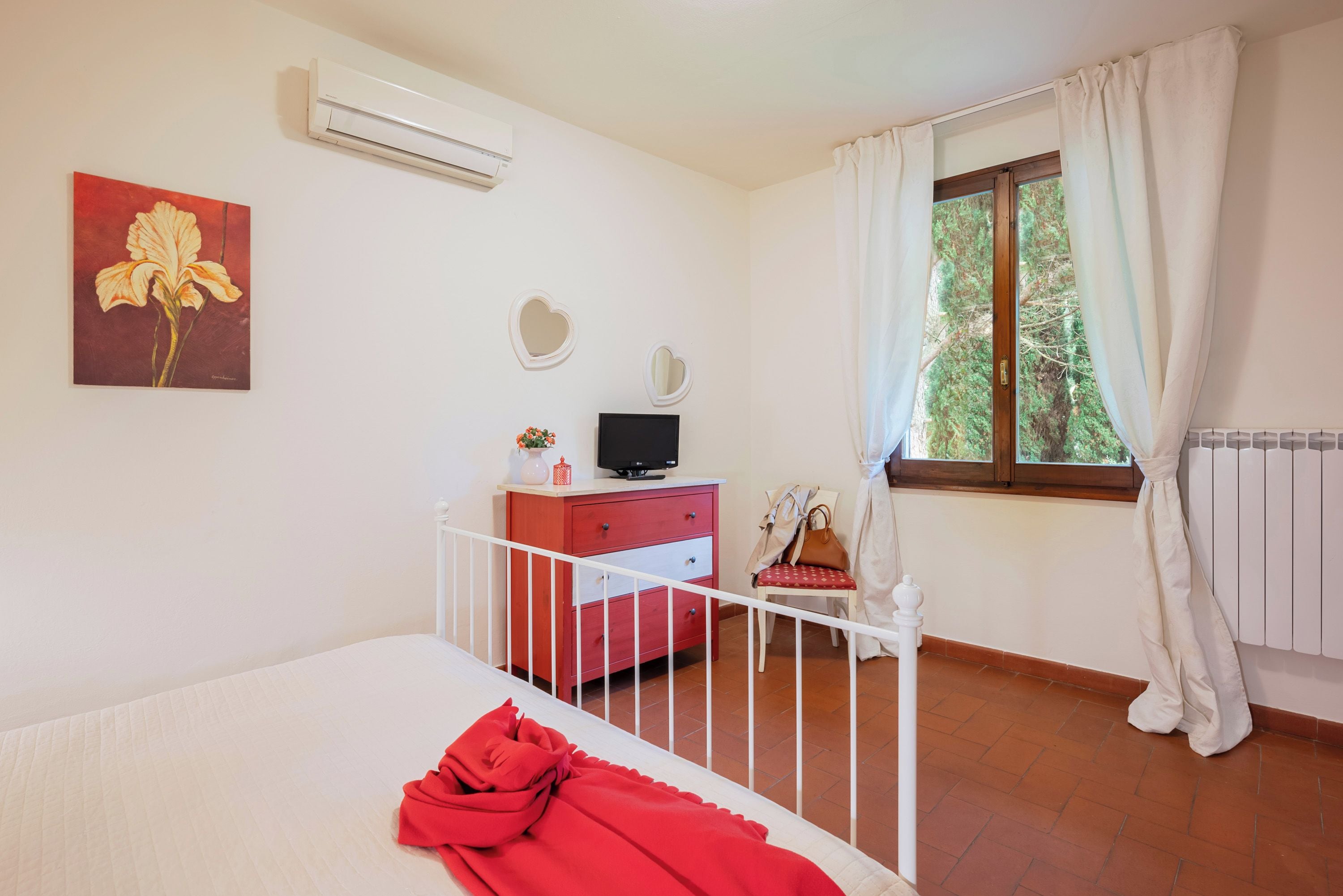 A bedroom with a bed, a dresser, a TV, a window with curtains, and a painting on the wall.