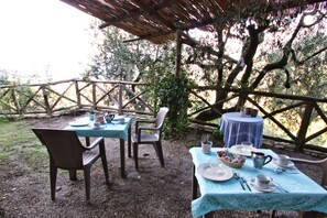 Zona pranzo all'aperto con tavoli e sedie, circondata da alberi e un recinto in legno.
