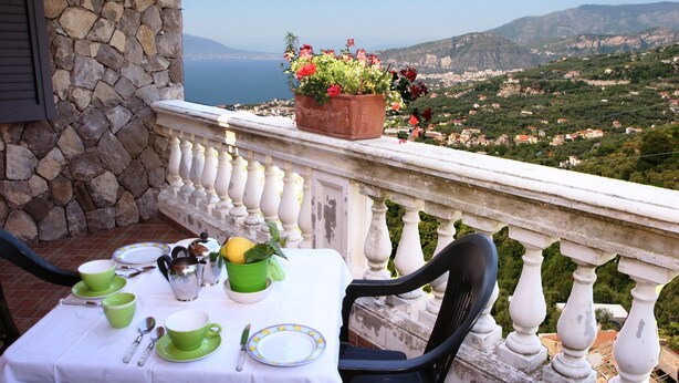 A terrace with a table set for tea, a potted plant, and a scenic view of a coastal town. - Casarufolo Paradise (Sorrento)