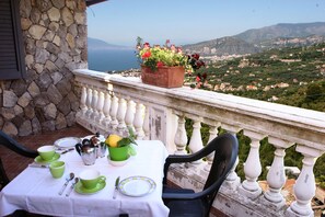 A terrace with a table set for tea, a potted plant, and a scenic view of a coastal town. - Casarufolo Paradise (Sorrento)