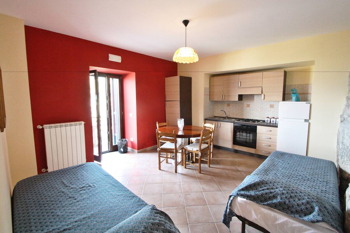 A compact room with two beds, a dining table with chairs, a kitchen area, and a radiator.