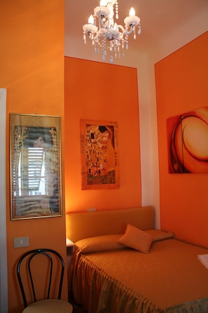 A room with an orange wall, a bed with a brown bedspread, a chandelier, and two paintings. - La Mimosa Bed and Breakfast (Lucca)