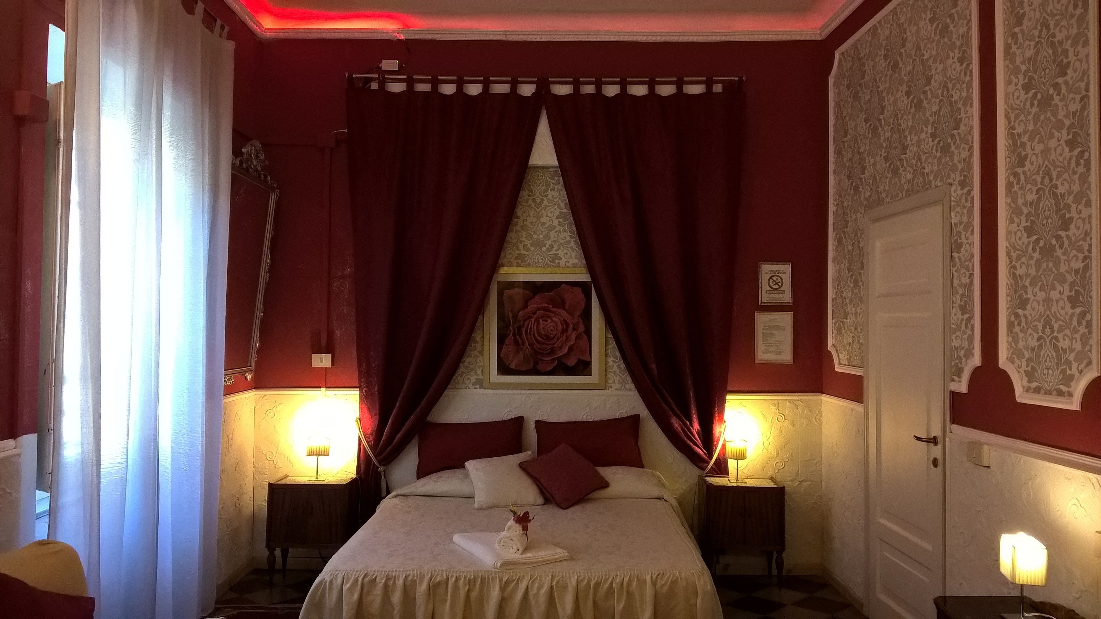 A hotel room with a large bed, red curtains, two bedside lamps, and a framed picture of a rose on the wall.