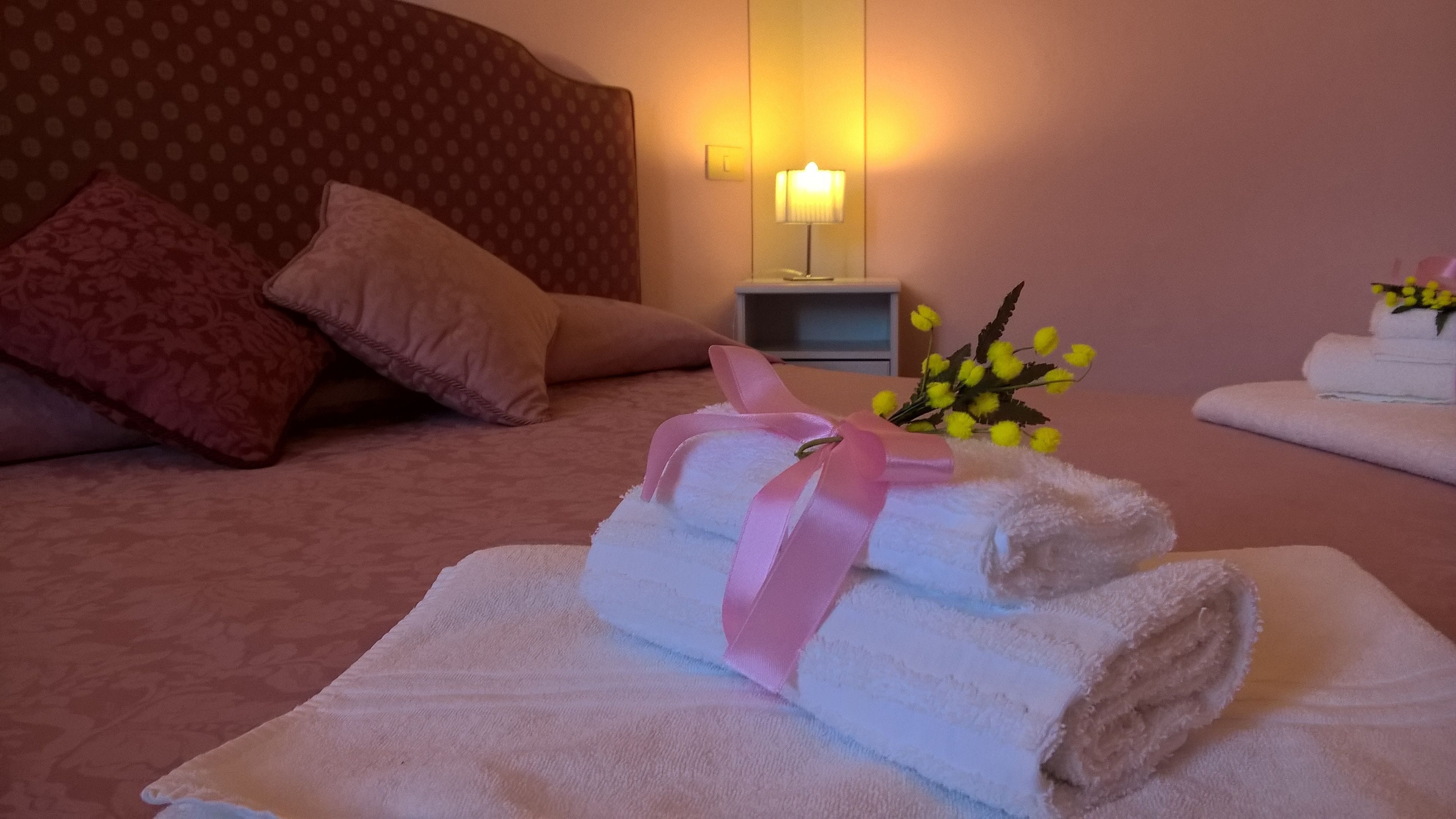 Un letto rifatto con cuscini rosa e beige, una lampada da comodino e un bouquet di fiori gialli.