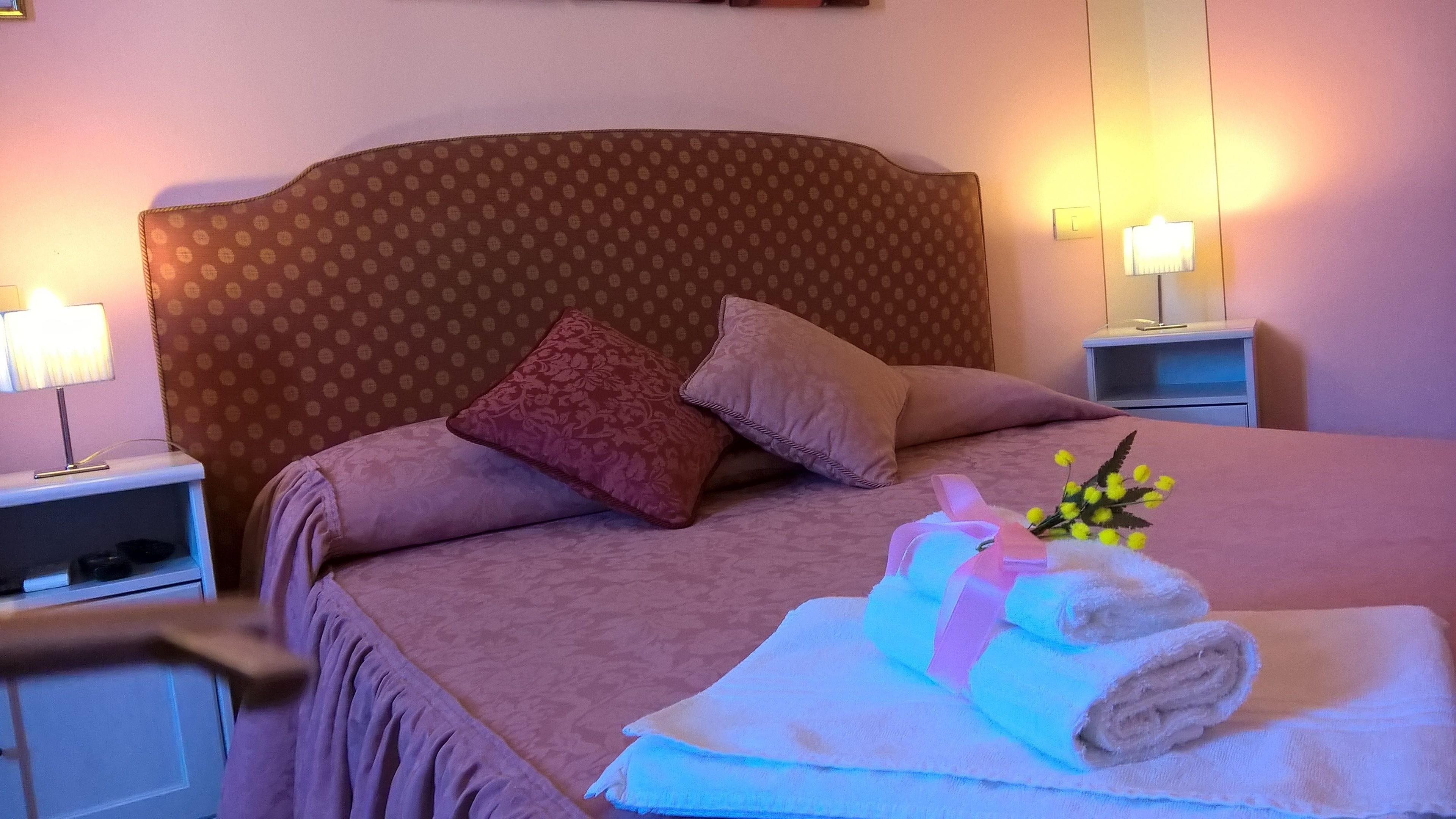 Uma cama bem arrumada com cabeceira estampada, duas almofadas, uma mesa de cabeceira com abajur e um criado-mudo.