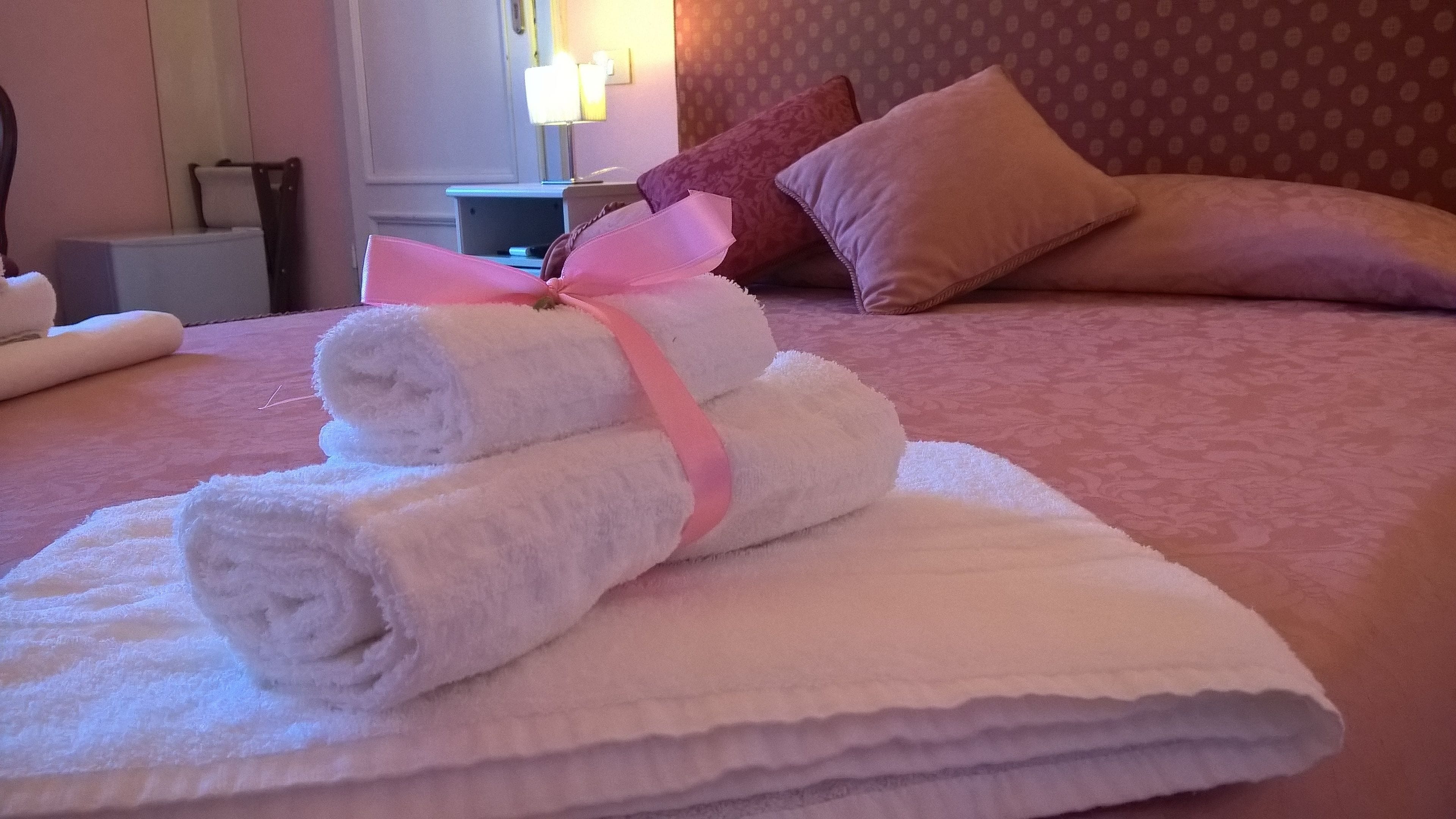 A neatly made bed with rolled white towels and a pink ribbon, a patterned wall, and a nightstand with a lamp.