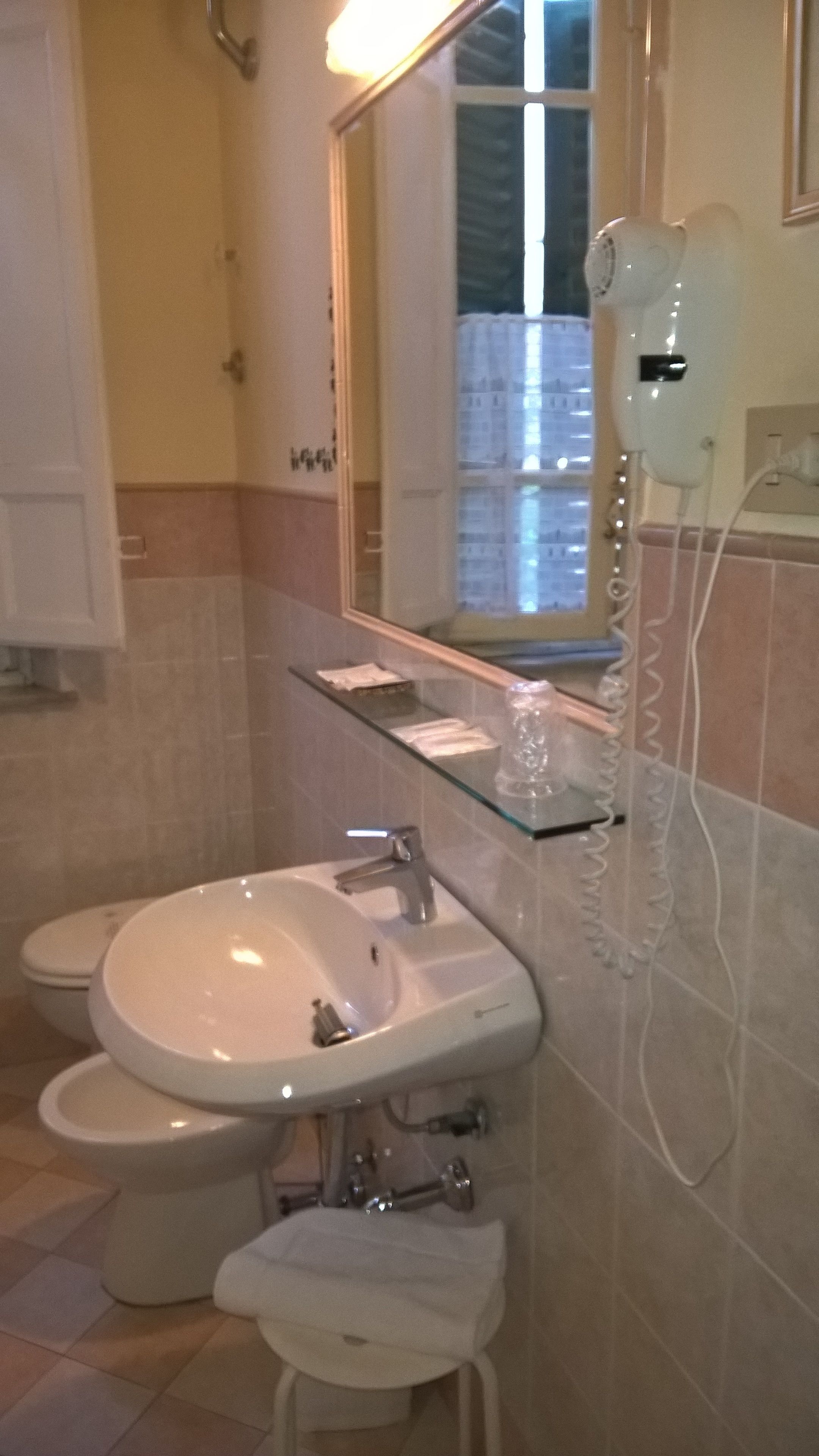 A bathroom with a white sink, two toilets, a glass shelf with toiletries, and a hairdryer on the wall.