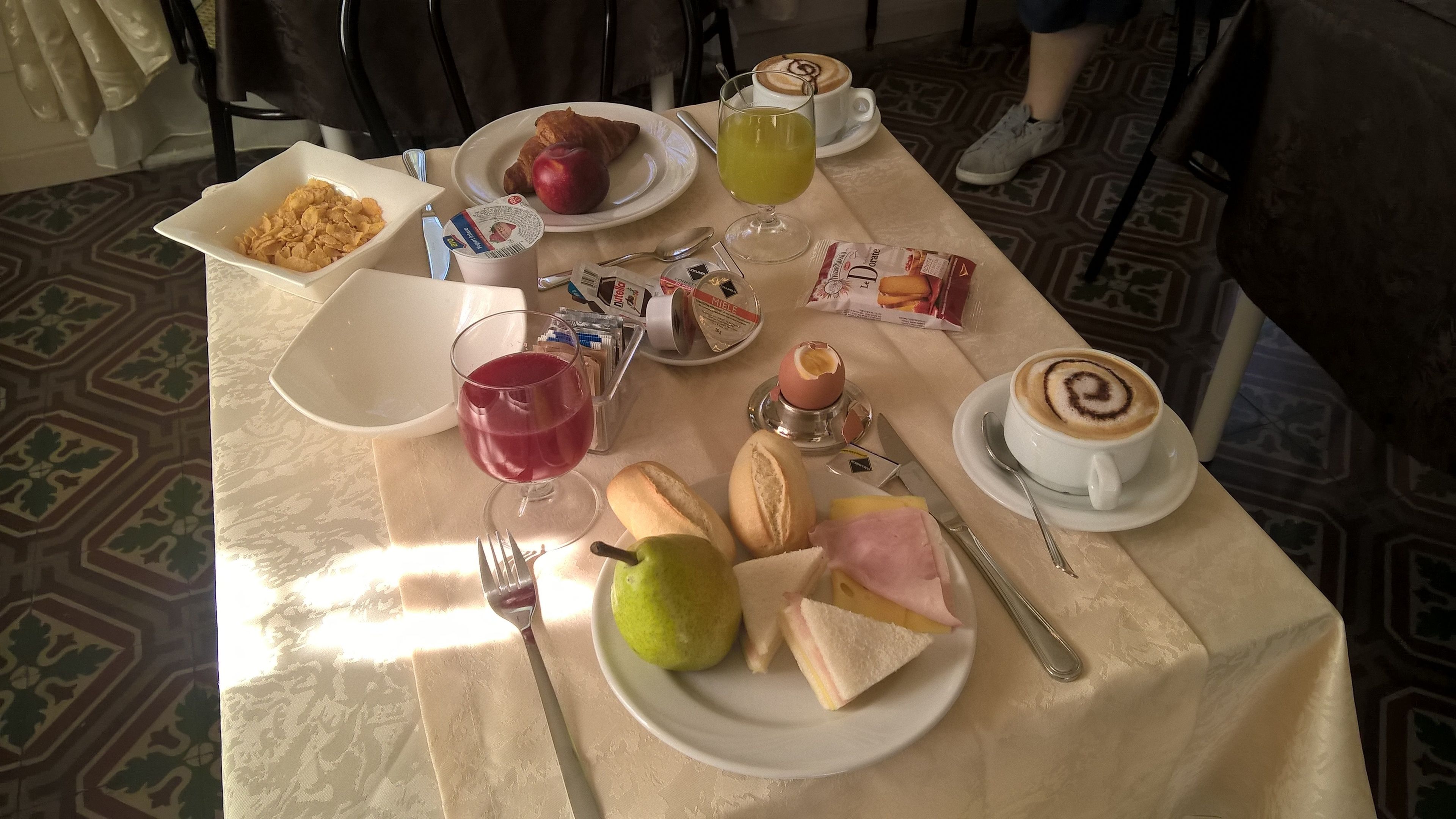A breakfast table with croissants, a pear, a ham sandwich, a glass of red juice, a glass of green juice, a cup of coffee with latte art, and a plate of cereal.