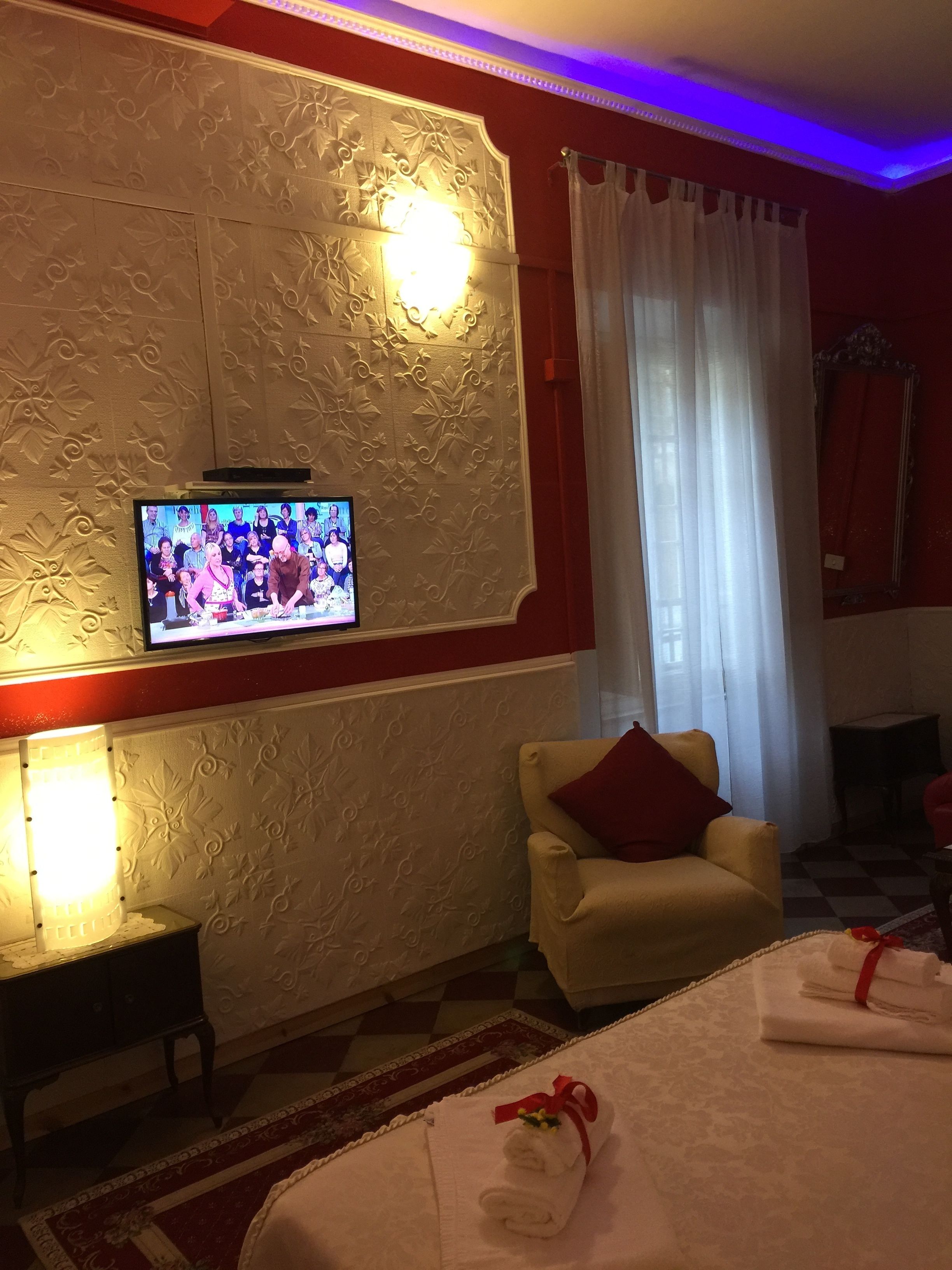 Une chambre d’hôtel avec un téléviseur fixé sur un mur décoratif, un canapé agrémenté d’un coussin rouge, une petite table et un lit bien fait avec des serviettes.