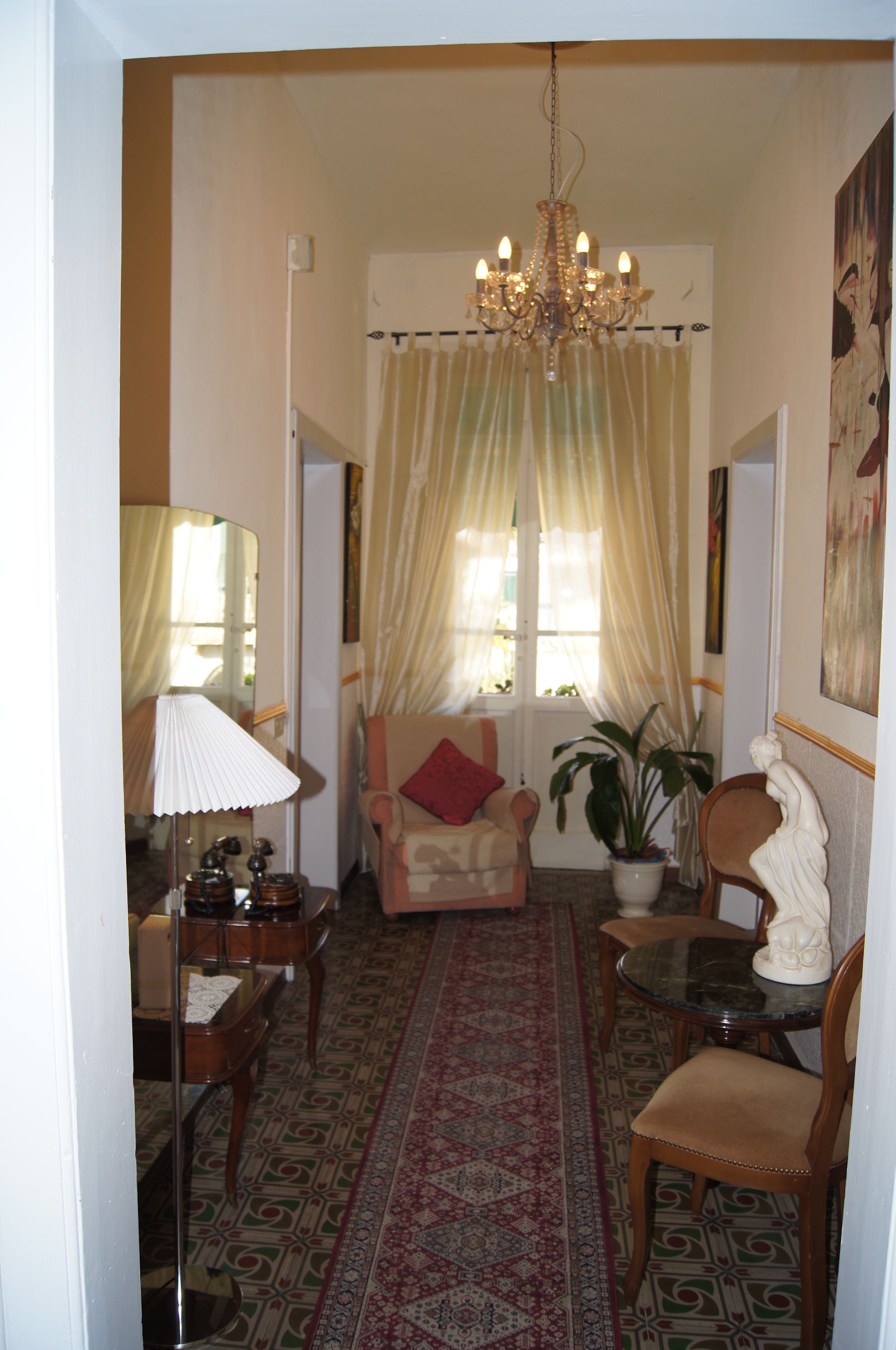 Un salon avec un lustre, un fauteuil orné d’un coussin rouge, une table avec une lampe et une plante en pot.