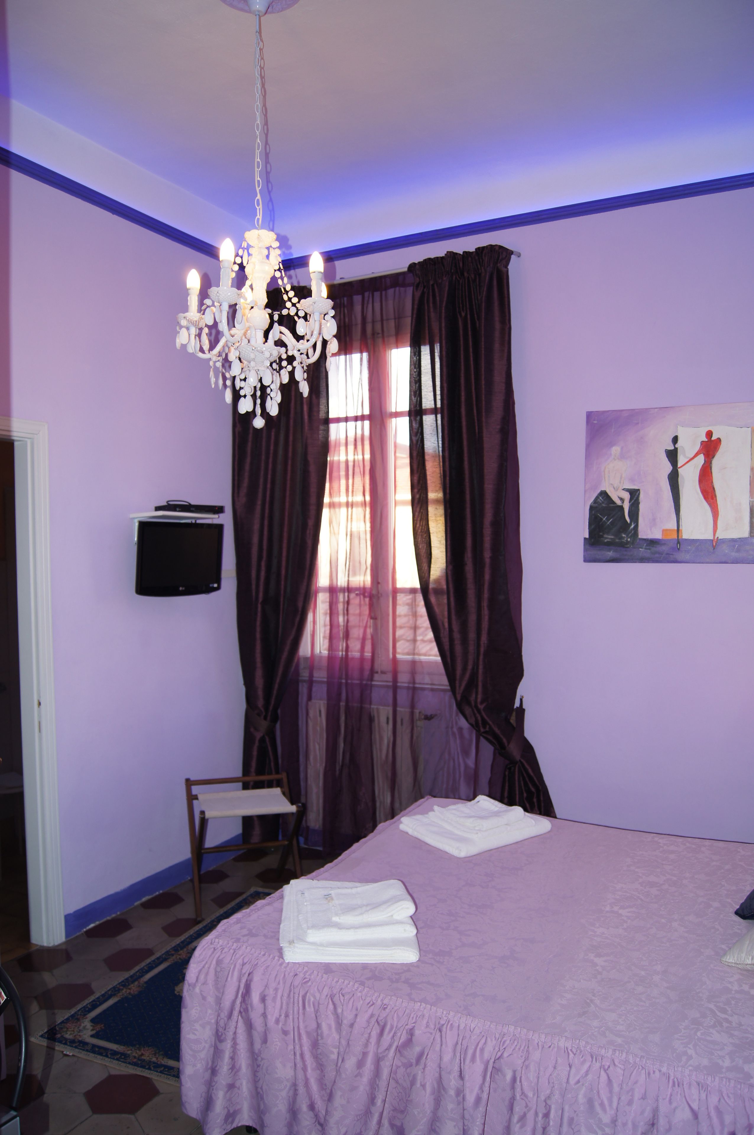 A bedroom with a bed, a television, a chandelier, a window with curtains, and a painting on the wall.