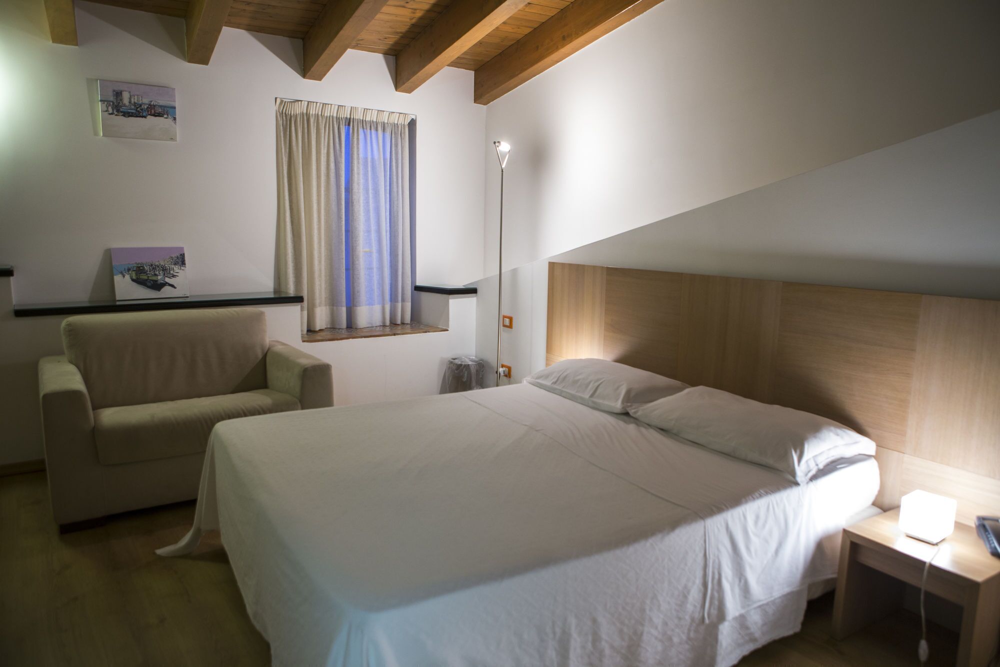 Un dormitorio con una cama, una silla, una mesita de noche, una lámpara y una ventana con cortinas.