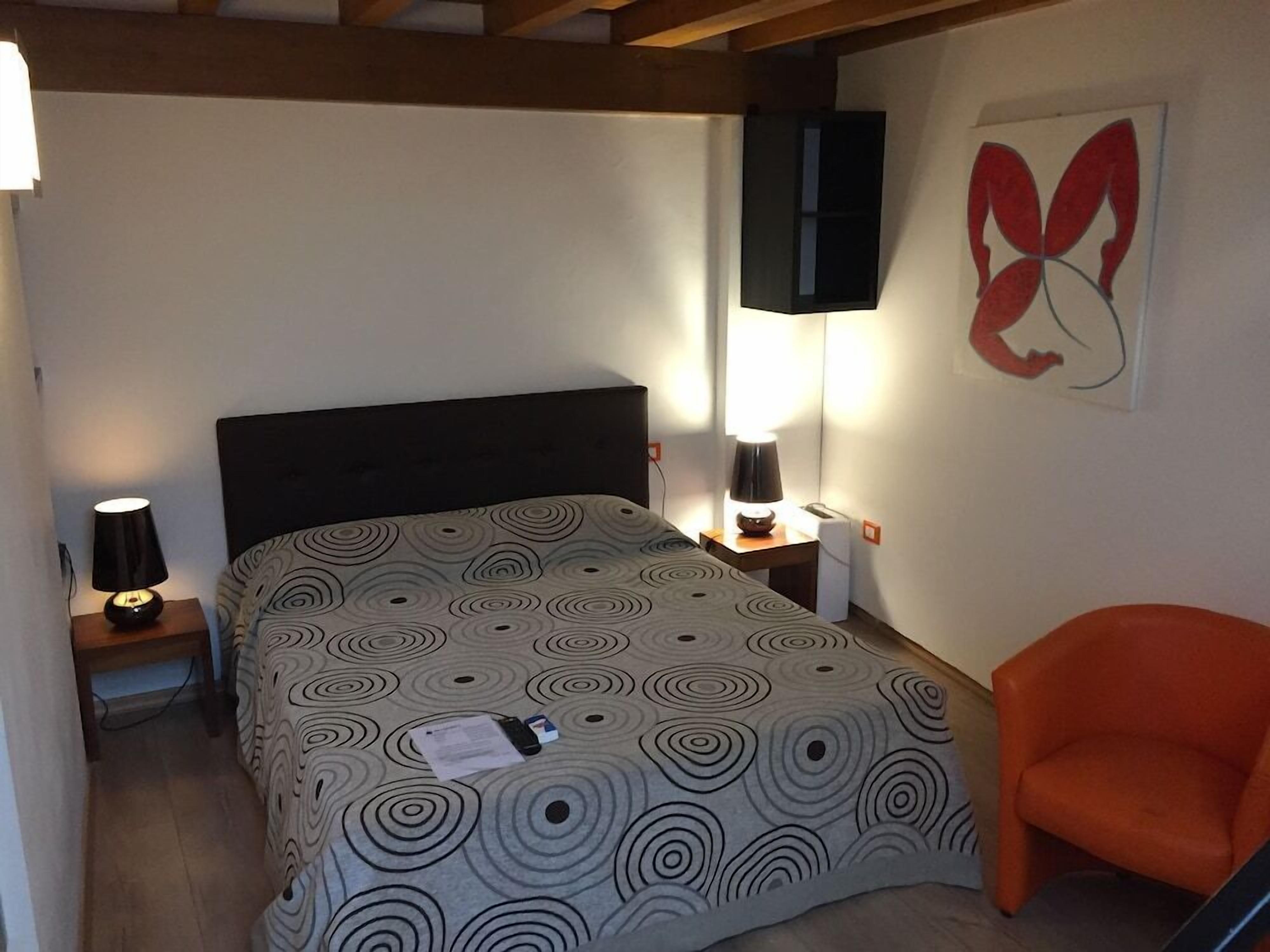 A bedroom with a bed, a nightstand, a lamp, a chair, and a wall art piece.