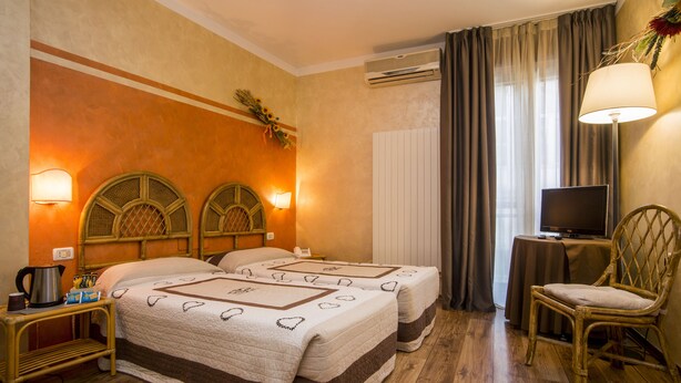 A hotel room with two beds, a television, a chair, a table with a lamp, and a wall-mounted air conditioner. - Le Pageot (Aosta)