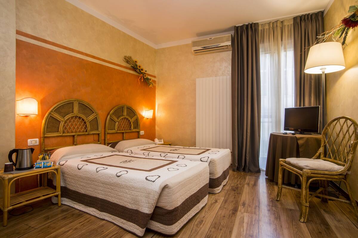 A hotel room with two beds, a television, a chair, a table with a lamp, and a wall-mounted air conditioner.