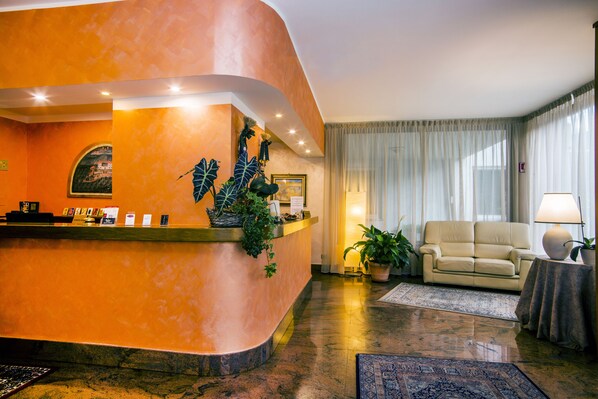 A hotel reception area with a curved orange wall, a reception desk, a beige sofa, and a potted plant. - Le Pageot (Aosta)