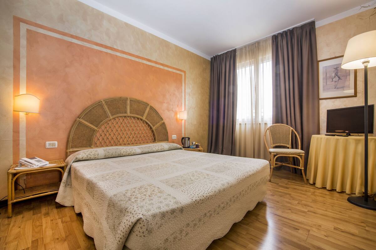 A hotel room with a large bed, a wicker headboard, a bedside table with a lamp, a chair, a TV on a stand, and a window with curtains.