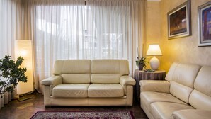 A living room with a beige sofa, a side table with a lamp, a potted plant, and a large window with sheer curtains. - Le Pageot (Aosta)